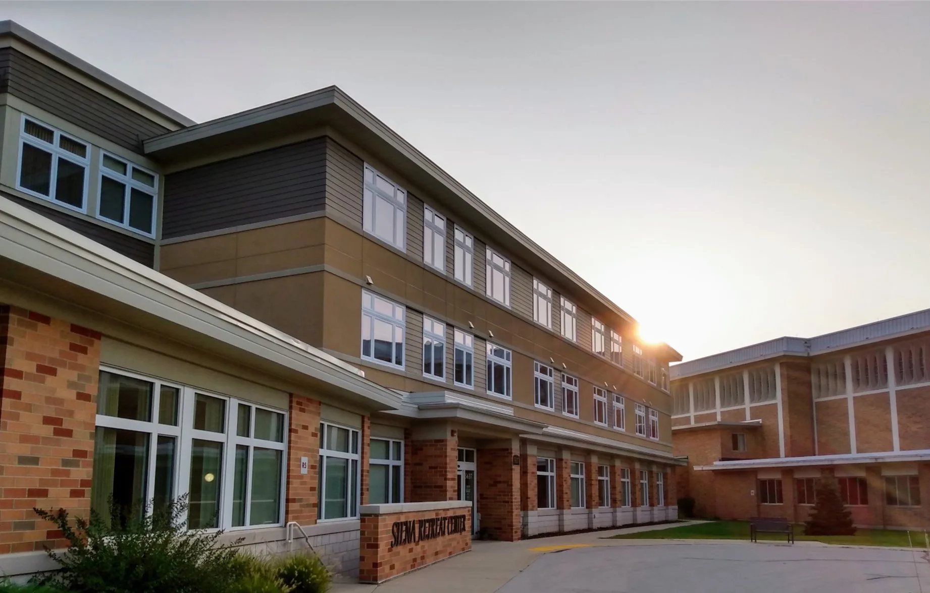 front of a three story modern building with sun in background and sign stating Siena Retreat Center